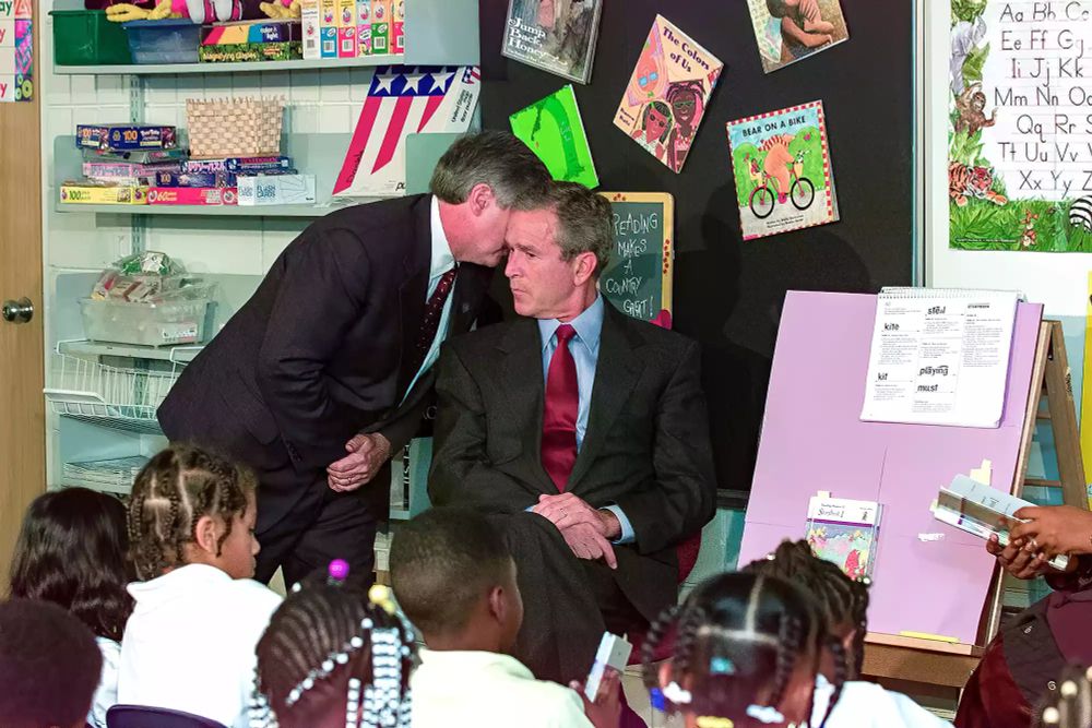 George W Bush vor Kindern, ein Berater flüstert ihm was ins rechte Ohr