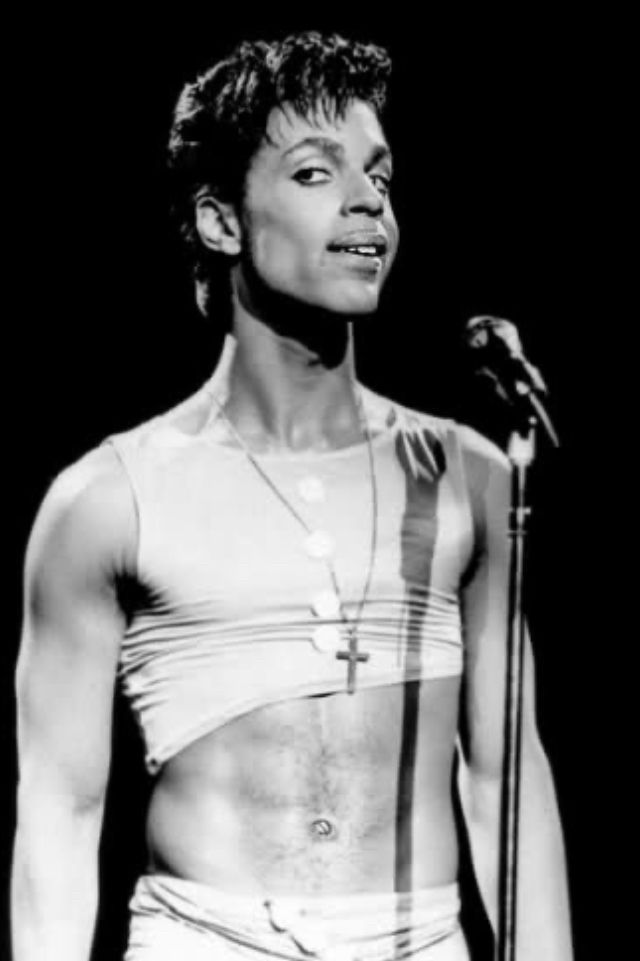 Black and white portrait of Prince at the microphone, his eyes looking askance, his body wearing crop top revealing 6 pack abs. 