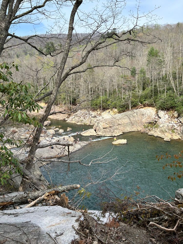 A central man in a yellow bathing suit stands on a rock surrounded by water near the edge of the basin at the bottom of Elk River Falls. Families with small children play in the shallow water at the edge of the river basin. The surrounding rock is scoured of leaves and moss. Tree trunks, and dead bushes are wedged at the sides of the trail. On the far side of the picture where the stone makes a corner of the basin, the metal frame of a vehicle trailer can be seen with the wheels intact, slightly mangled from where it had washed over the falls.