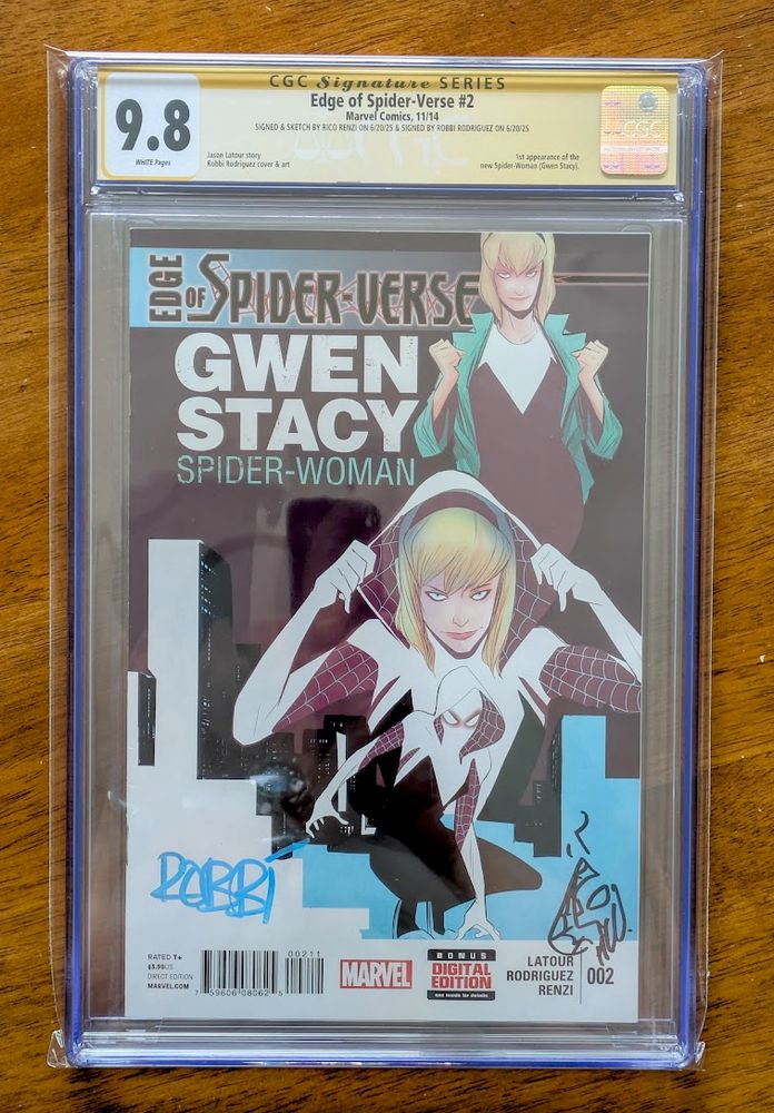 A photograph of a professionally graded comic book on a wood table. The comic book is Edge of Spider-Verse #2. The cover features three poses of Gwen Stacy as Spider-Woman/Spider-Gwen: standing upright holding the lapels of a jacket open, pulling her hood back to reveal her face, and squatting in full costume. The cover also has signatures from Robbi Rodriguez and Rico Renzi. The comic book is encased in a clear plastic slab, including a yellow label from CGC with a 9.8 assigned grade.