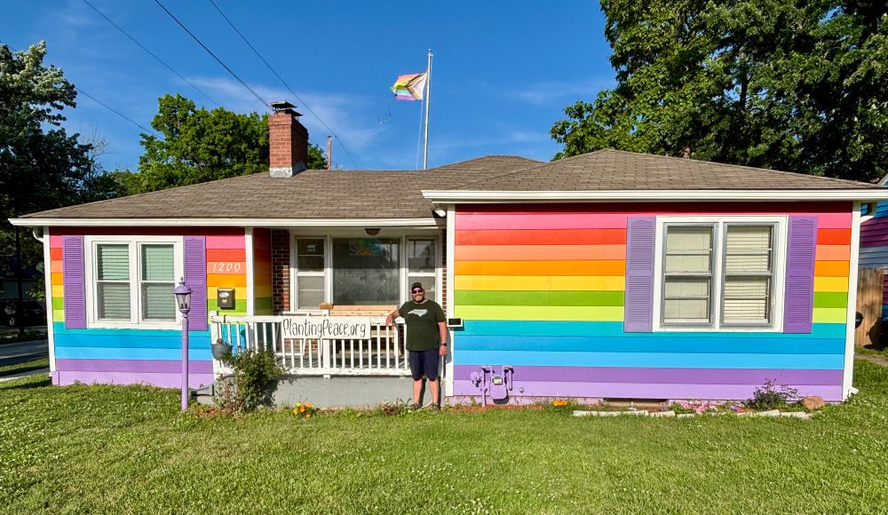Man standing in front of a house painted in rainbow colors of the Pride Flag. Green grass in foreground. Plantingpeace.org shown on a sign 