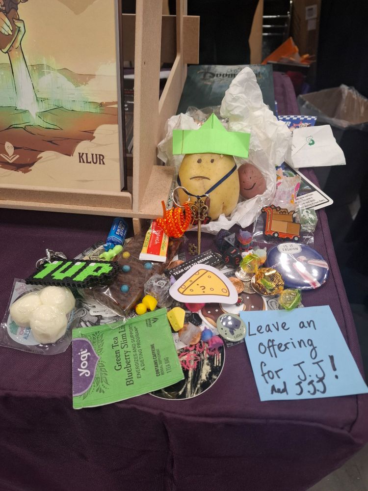 A potato with a sad face, green paper hat and key surrounded by various trinkets and offerings with a post it note sign that reads "leave an offering for JJ"