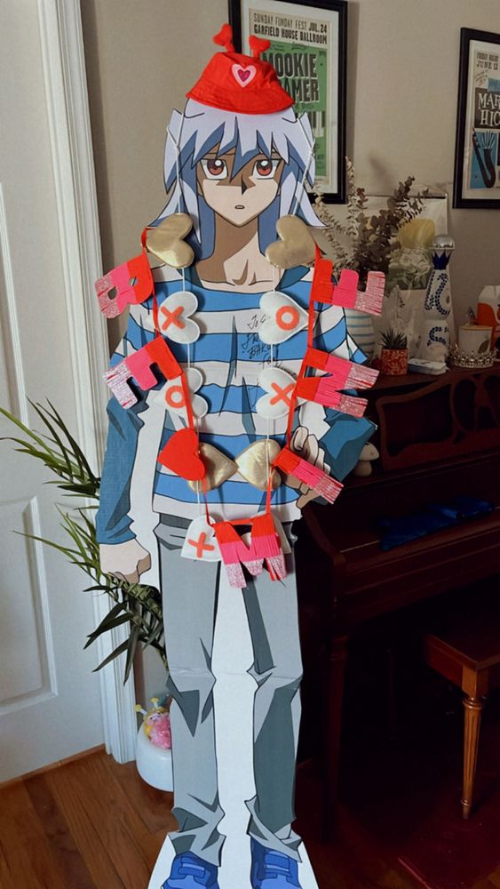pictured is a canon height cardboard cut out of YuGiOh character, Ryou Bakura in the DSOD art style wearing his blue and white striped shirt, navy undershirt, gray jeans, and blue sneakers. he is decorated for valentine's day! there is a pink and red garland around him that says Be Mine and a pink, white, and gold garland that has X's and O's. on top of his head he's wearing a little head hat with a pink heart and two red heart antennae sticking out!
