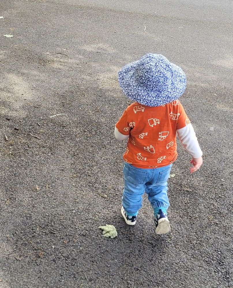 A photograph of a toddler taken from behind. His face and hair are hidden from view by a big blue and white floral hat. He is wearing a bright orange t-shirt with white doodles of vehicles, a white long-sleeved vest underneath, blue jeans and navy and white trainers. He is walking on gravel.