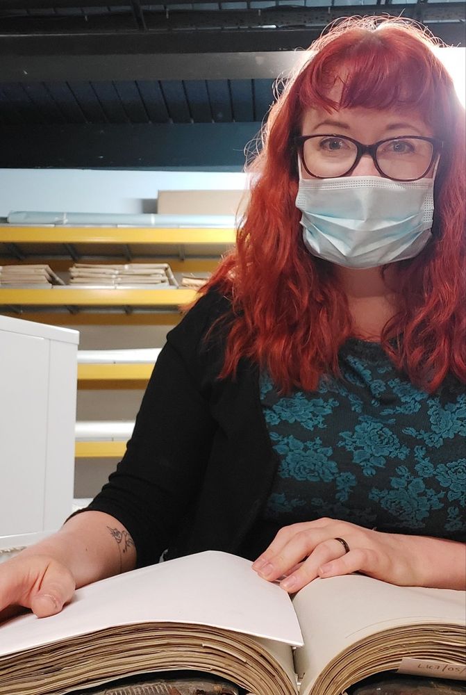 A woman with red mid length hair and a fringe, wearing a disposable pale blue mask and dark-rimmed glasses, a black dress with turquoise flowers, and a black cardigan. She is seated at a table with an historical book open to a page. The identifying information of anyone on that page has been covered by a sheet of paper for data protection purposes. Behind the woman are a series of shelving units with archival documents.