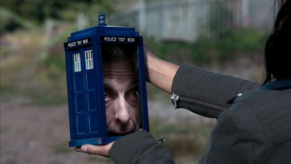A small deep blue police telephone box held in a woman's hands, with the doors open and Peter Capaldi's face filling up the doorway.