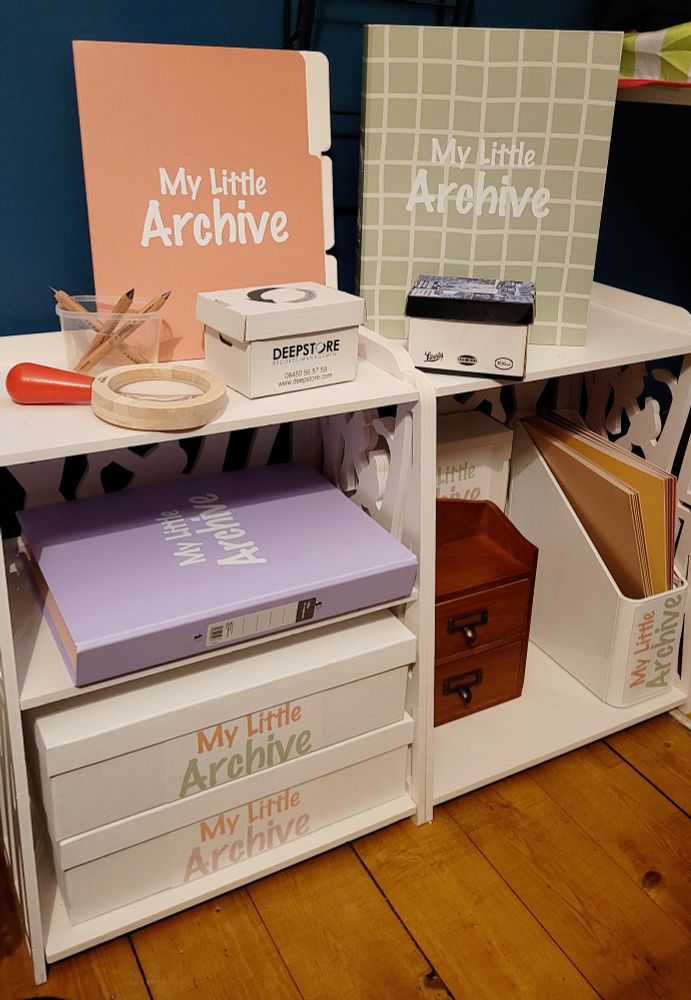 Two small white bookcases with boxes, drawers and folders inside them. In the bookcase on the left, there are two white card boxes with the words "My Little Archive" in pastel font colour. There is also a lilac ring binder with "My Little Archive" in white. On the top of the left bookcase is a small kid's wooden magnifying glass with a red handle, a small version of a box from DeepStore, a clear plastic tub with pencils, and a peach coloured file that says "My Little Archive" on in white text. In the right bookcase, there is a small wooden card catalogue with two drawers, a white magazine file with files inside, and on the top of the bookcase is a green ring binder with white horizontal and vertical lines in a criss-cross pattern, and the words "My Little Archive" in white. Next to this is another small box like a model Bankers Box.