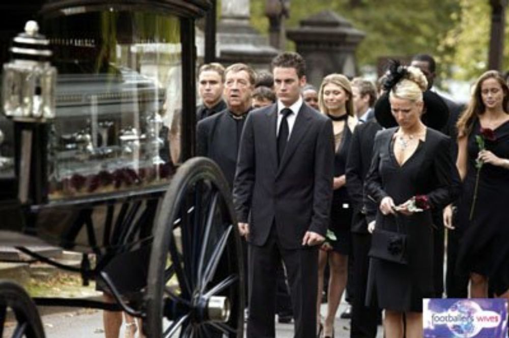 A funeral. A black horse-drawn carriage with a silver coffin inside. Behind the carriage are mourners dressed in smart black clothes. They are the characters of Footballers Wives.