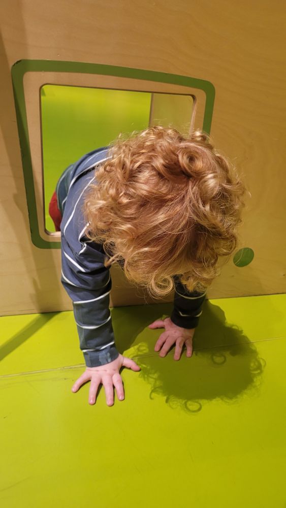 A photograph of a toddler with strawberry blonde curly hair climbing out of a square hole cut into a wooden panel at a kid's play area in a museum. He is wearing a navy long-sleeved top with white horizontal stripes. His head is face down and you can only see the top of his head. His palms are outstretched on the lime green floor