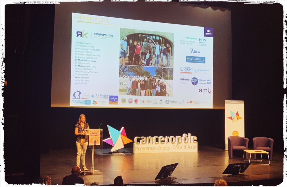 Marion Le Grand standing on the stage of the Canceropole PACA annual congress, in front of a huge screen showing her final slide, with 2 team photos, the names of team members and collaborators, as well as logos of participating research institutions and funding bodies.