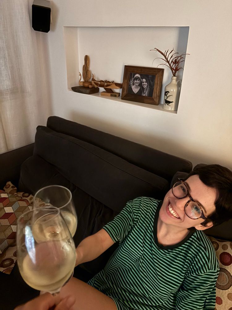mulher branca com uma taça de vinho verde na mão brindando com a taça que eu, que tirei a foto, seguro