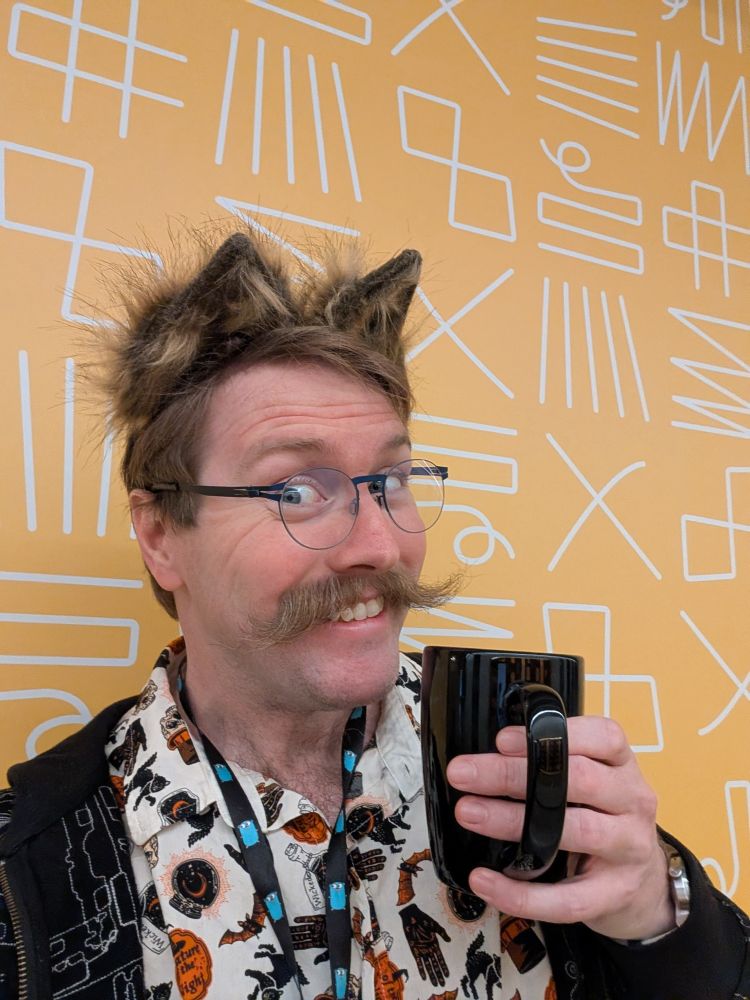 A perfectly ordinary selfie of Robert in front of a yellow background with abstract lines on it. He's holding a coffee mug, and grinning at the camera. He's wearing a Halloween themed shirt, a black patterned hoodie, and fox ears that match his hair.