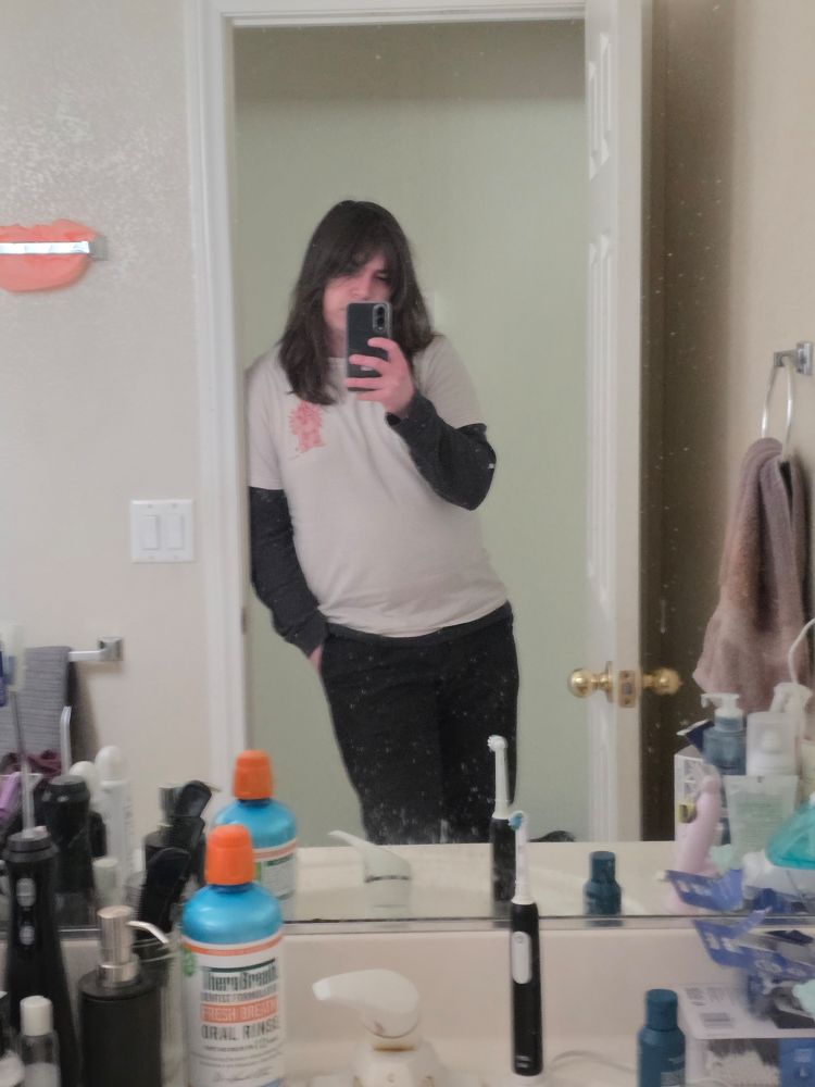 A bathroom mirror of selfie of Burtch wearing a white t-shirt with a thermal underneath and black pants. Burtch's phone blocks part of their face.