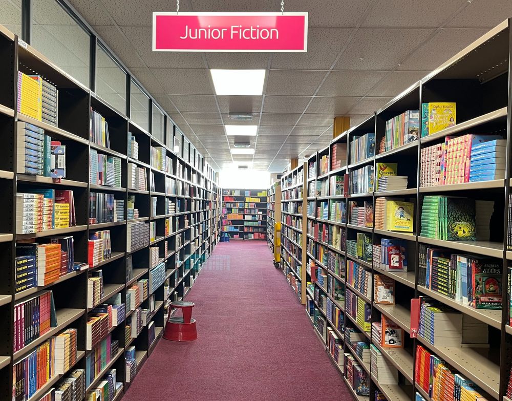 Colour image of two long rows of tall bookshelves. Hanging from the ceiling in the top centre of the image is a pink sign saying 'Junior Fiction'. At the end of the row are more bookshelves, lined with colourful books.