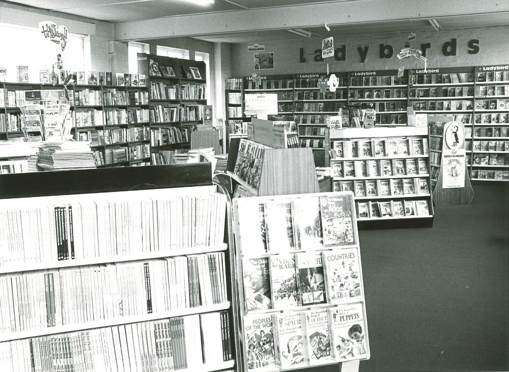 Black and white image of Peters bookshop from the early 1980s. At the front of the image, there is an arrangement of short bookcases stacked with titles. In the background, there is tall shelving with Ladybird books. Illustration mobiles hang from the ceiling.