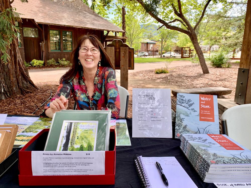 Smiling woman at outdoor table selling haiku books, prints and cards. Photo taken at ukiaHaiku Festival 2022 in Ukiah, California.