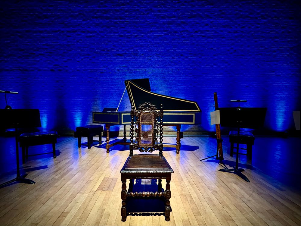 A chair on a stage in front of a harpsichord, blue lit background.