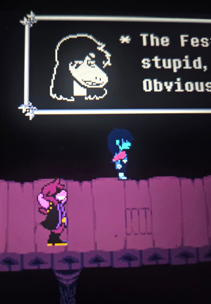 A photo of Deltarune Chapter 3 on my Steam Deck. It features Susie and Kris in the Dark World. Susie is seen speaking, but the text box is cut off so you can only read:
"The Fes
stupid,
Obvious"