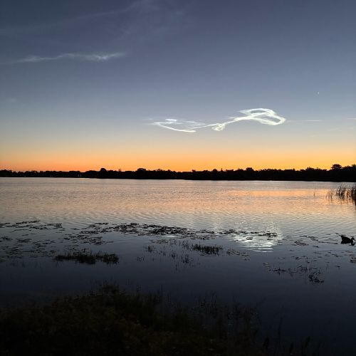 dawn's golden orange hues on the horizon and a clear sky with the loop-de-loop remains of rocket launch exhaust floating above the calm lake water