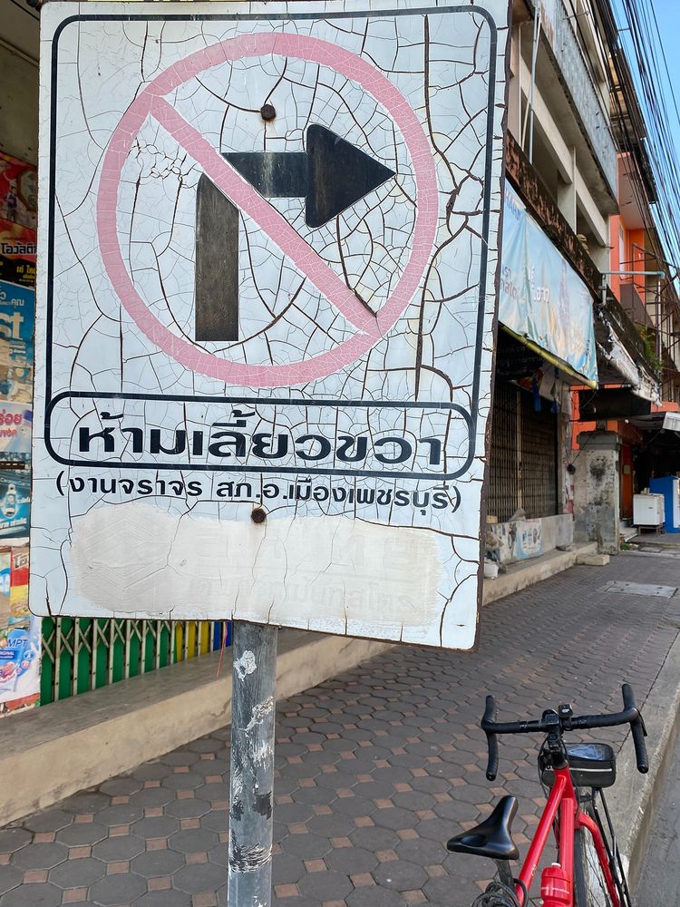 Photograph of a no-right turn sign beside a road in Petchaburi, Thailand. The sign is old with a network of fine cracks across it but is still reasonably legible. Behind the sign is a pavement (sidewalk) with closed shophouses plus my red bicycle.