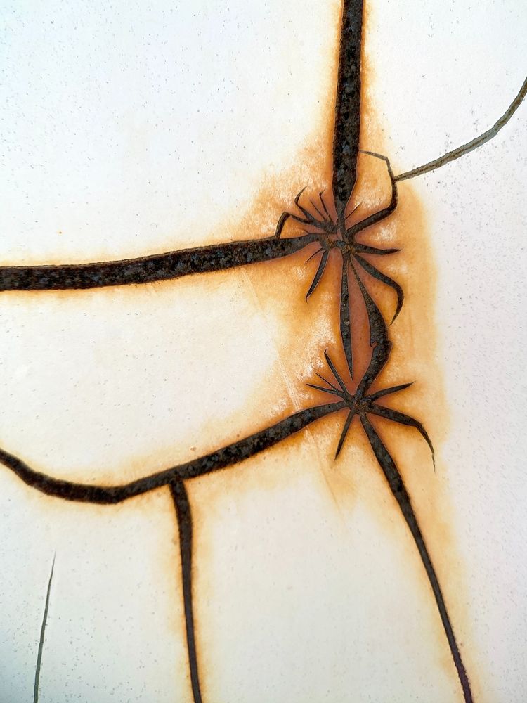 Close-up detail of cracked paintwork on the back of a road sign in Thailand. The cracks show the dark rust underneath and the white paint has been stained orange along the edges of the cracks. Two small star-like crack patterns resemble spiderlings in a corner of a large web.
