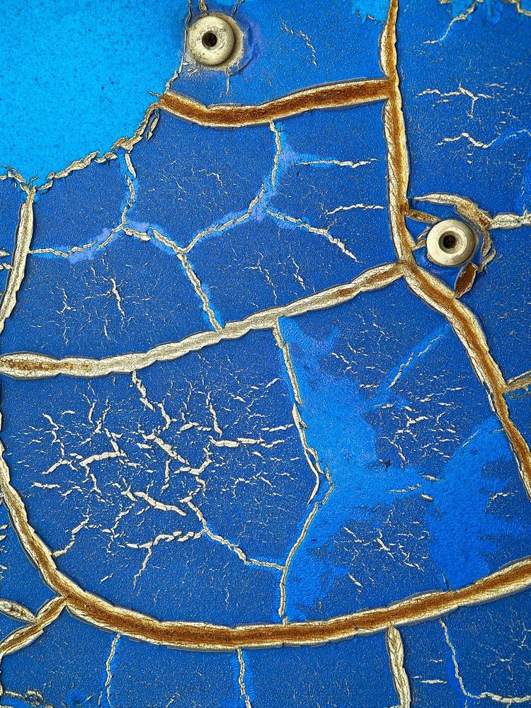 Close-up detail of cracked blue paintwork on the front of a road sign in Thailand. The cracks are silvery with a hint of dark rusting and two rivets combine with a curved crack to resemble a face.