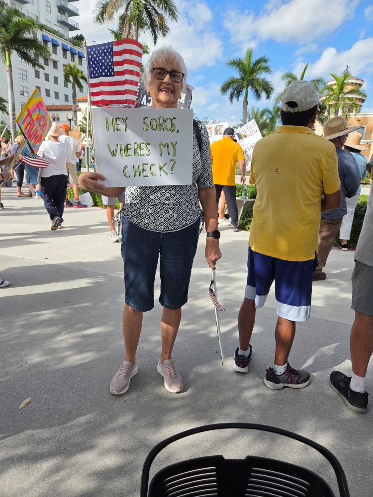 Elderly woman holding protest sign which says " Hey Soros, where's my check?" referring to claims by right wing politicians who claim George Soros is paying protesters!
