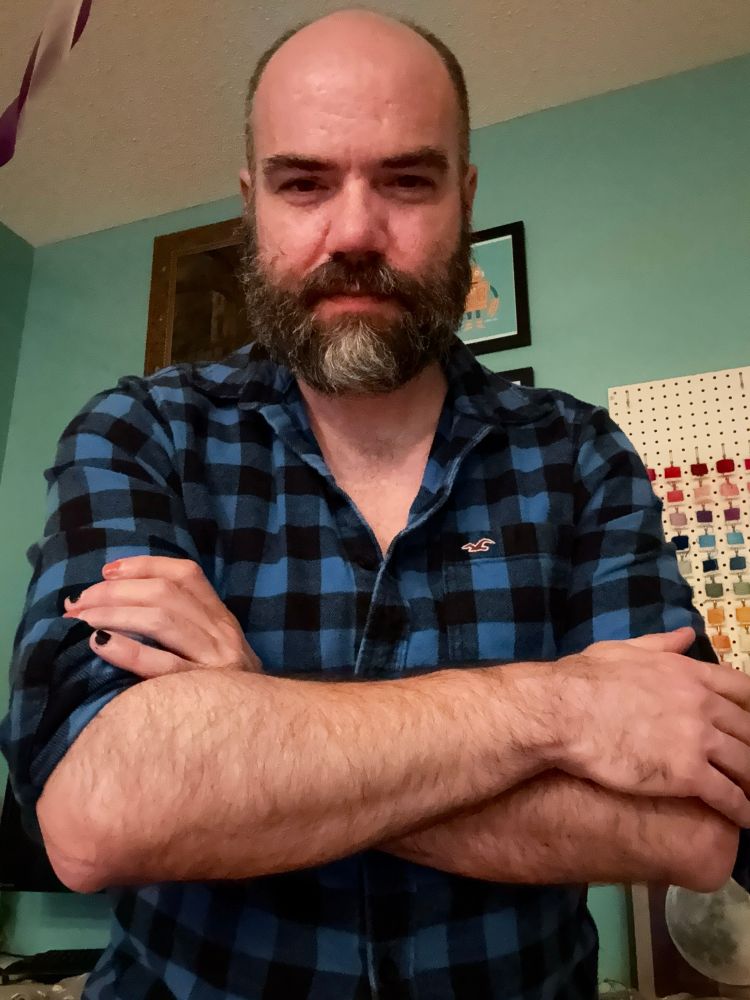 A picture of me, a white man with a brown beard and a mostly bald head, folding my arms with my blue and black flannel shirt’s sleeves rolled up 