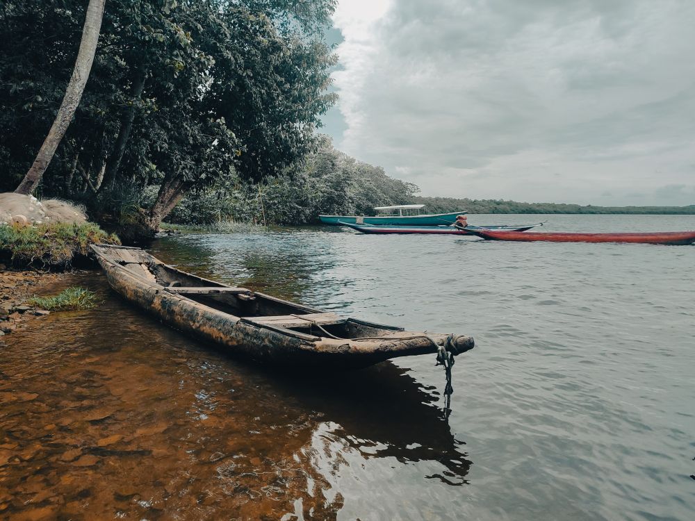 Foto de paleta de cores frias da beira do rio Jaguaripe em Maragogipinho, com sua água escura e calma com alguns pequenos barcos flutuando. O barco que está em destaque em primeiro plano é de madeira antiga e desgastada com uma corda pendurada na proa. Vê-se árvores e vegetações exuberantes na margem.
Na paisagem vê-se o céu nublado acima do rio.
Em segundo plano, 3 barcos coloridos contrastam com o barco de antigo.
