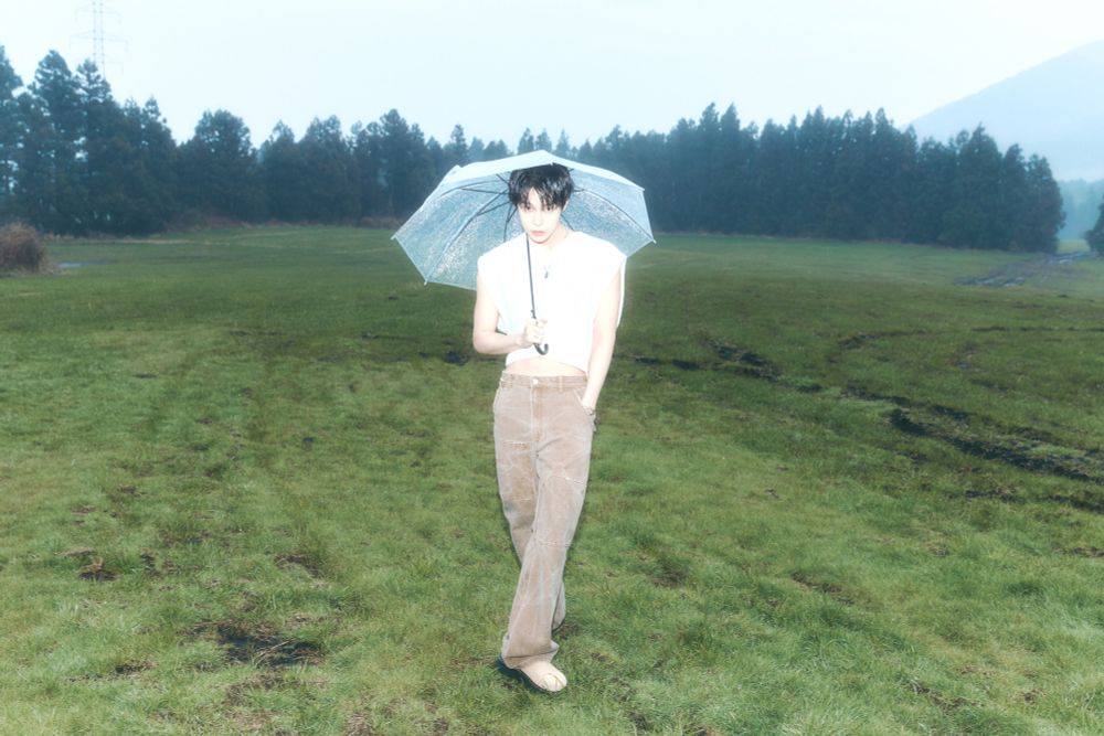 a photo that has the exposure all the way up making the lighting on doyoung superbly white and pale. he's standing with a clear umbrella over his head, one foot crossed over the other. he's also wearing a cropped, sleeveless white sweater and beige pants. the other hand is in his pocket and he's looking right at the camera.