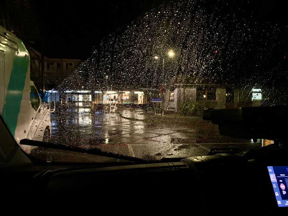 View through a rain-covered vehicle windscreen of a low, half-lit public building.