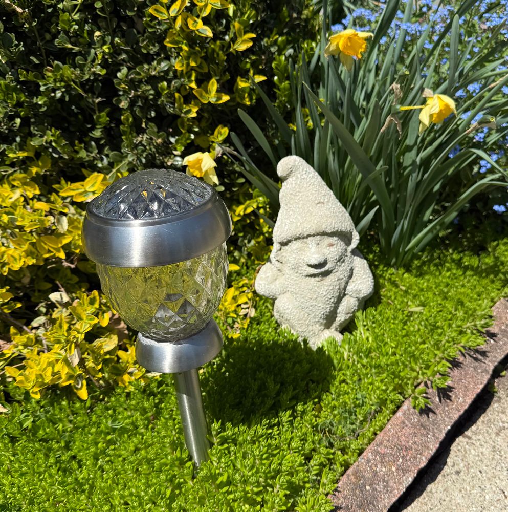 Adorable stone gnome sits in a flowerbed next to a solar walkway light.