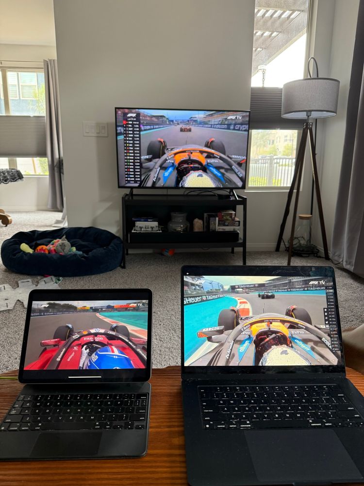 F1 main feed on big tv. Lando Norris feed on laptop on right. Charles Leclerc feed on iPad on left. iPad and laptop on wooden bench positioned in front of tv
