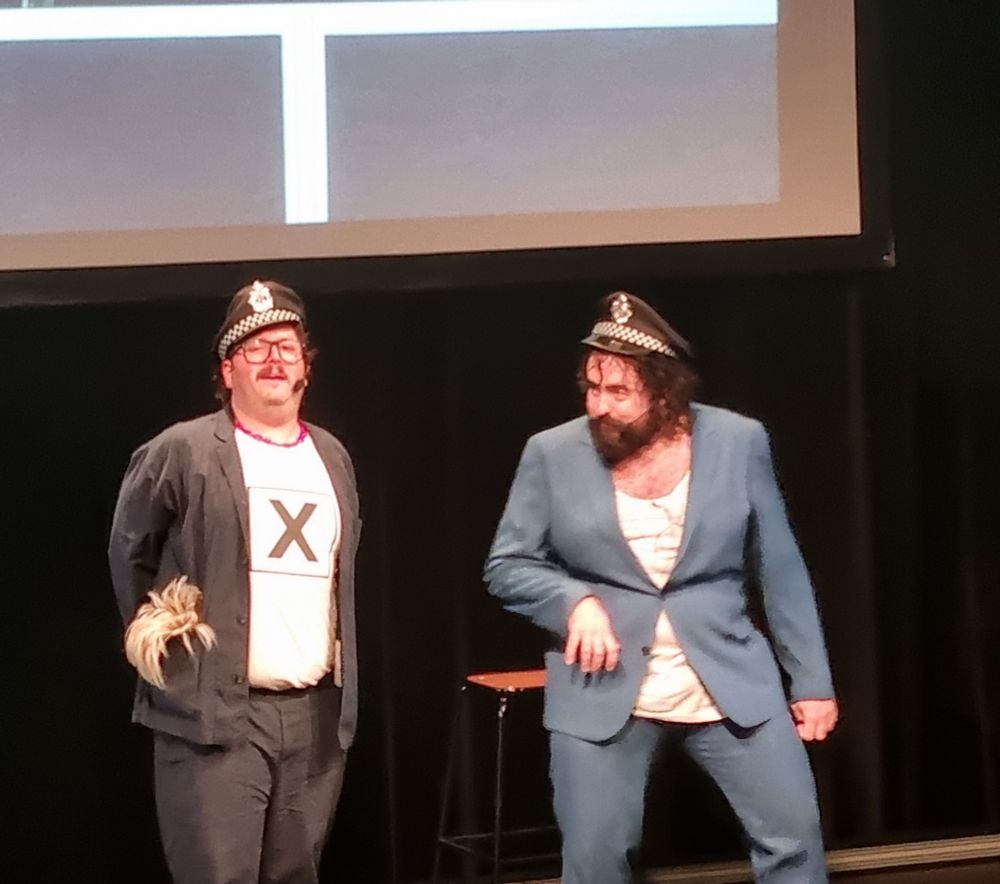 Two men on a stage. One wearing a police hat with a white shirt that has a big X on it and a regular jacket. The other wearing a blue suit and a police hat.