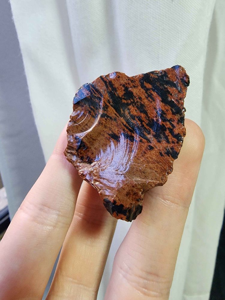 A Mahogany Obsidian being held up, it's red with black streaks. It looks smooth but the reflection of the light reveals it's curves.