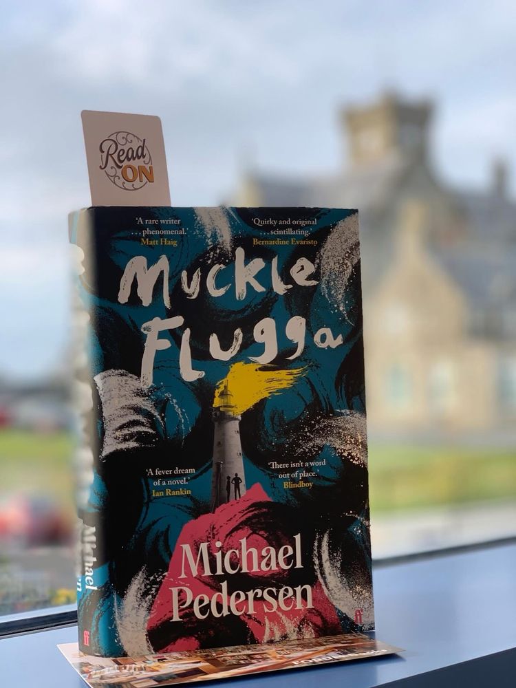 Muckle Flugga book in library window, book cover is bright design in black, white, red, blue and yellow.
