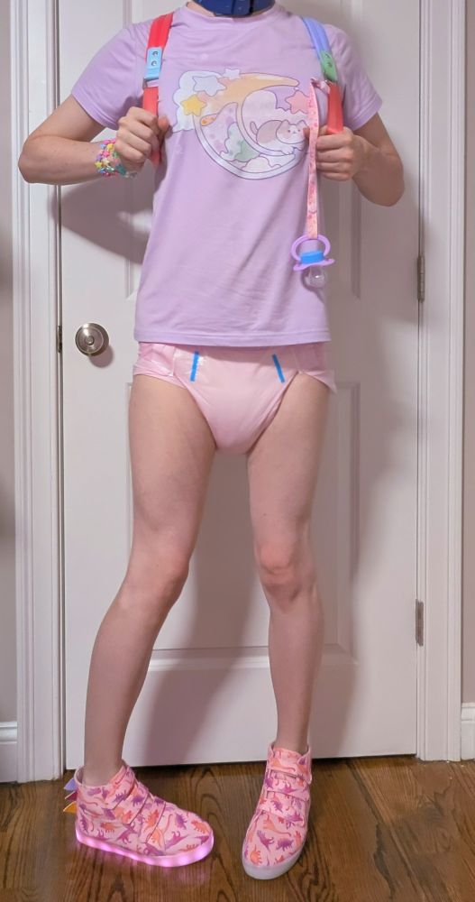 A person in a purple shirt, a pink diaper and light up pink shoes, holding onto their backpack straps