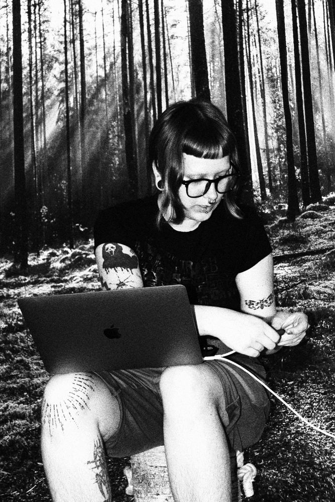 A black-and-white photograph of a person sitting against a wall giving an illusion that they are in a forest But it is just a picture wall, using a laptop. The individual has tattoos on their arms and legs, wears glasses, and has short hair. The forest background is dense with tall trees.