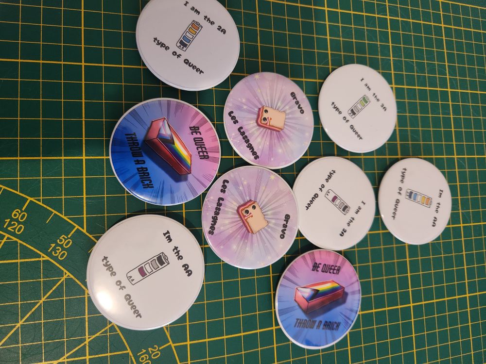 Differents badges avec des illustrations queer dessus :
Bravo les lasagnes (av3c un dessin de lasagnes lesbiennes)
Be queer throw a brick (avec une brique aux couleurs du drapeau arc en ciel progressif)
I'm the AA/2A type of queer (av3c des piles aux couleurs du drapeau aroace ou aro ou ace)