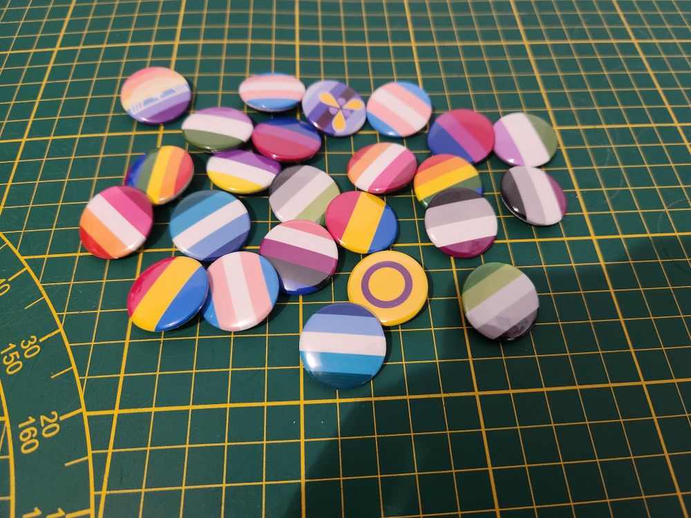 Pleins de petits badges aux couleurs de différents drapeaux LGBTQIA :
Lesbien, gay, bi, trans, intersexe, genderqueer, aro, ace, pan, xenogenre