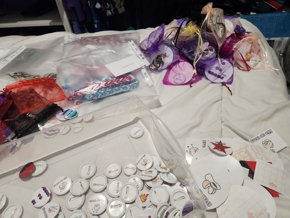 Photo du bout d’un lit on y voit pêle-mêle :
Un tas de petites pochettes en oeganzas de différentes couleurs, remplies de stickers badges et bracelets.
Un tiroir en plastique avec des badges dedans
Un tas de stickers 
Un tas de pochettes vides et des pochettes plastiques contenant des bracelets