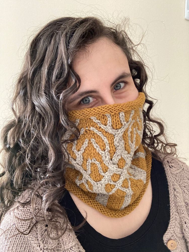 A woman is wearing a cowl of mustard yellow. On the cowl are cables that create a forest motif. The cables are silver. The woman has brown hair and is looking at the camera. Her nose and below are hidden by the cowl.