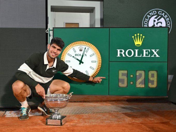 Carlos Alcaraz posing with the French Open men's trophy, pointing at a clock that says "5:29"