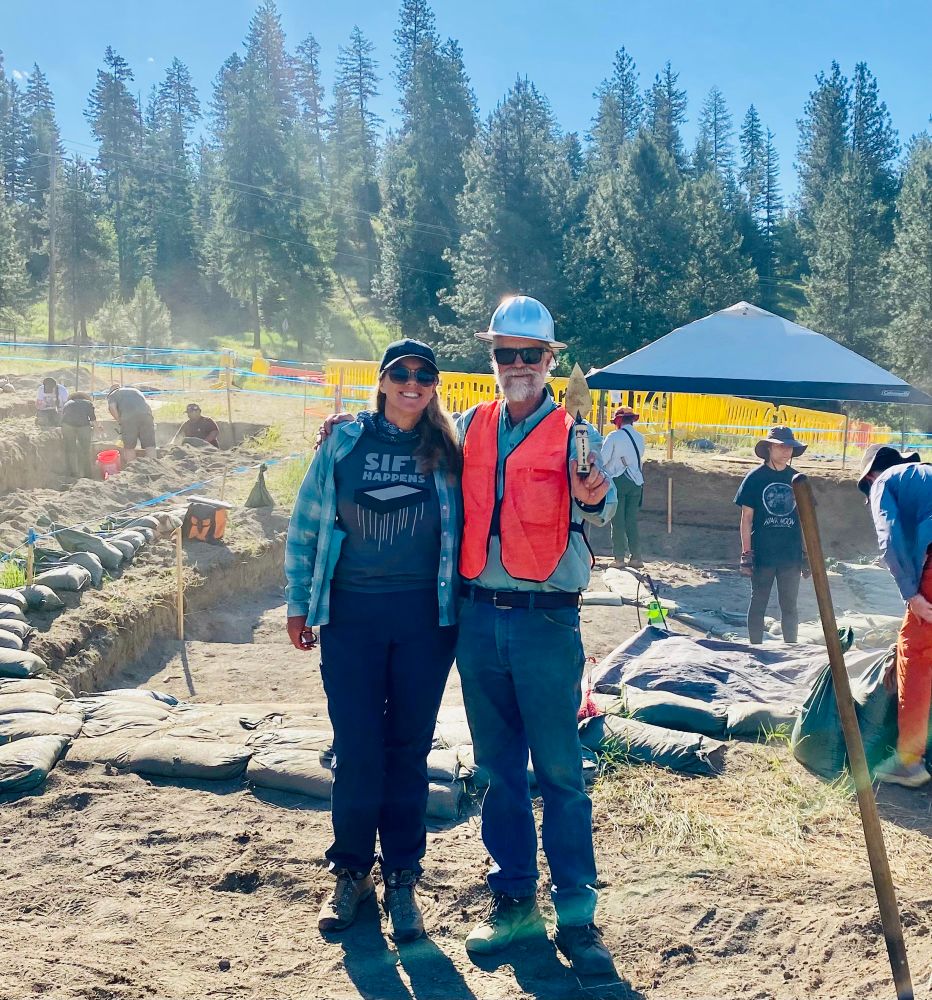 Shannon Tushingham and Bill Hildebrant smiling  in front of an archaeological excavation with students working. Shannon is wearing a T-shirt that says happens and Bill is wearing a hard hat and holding a trowel