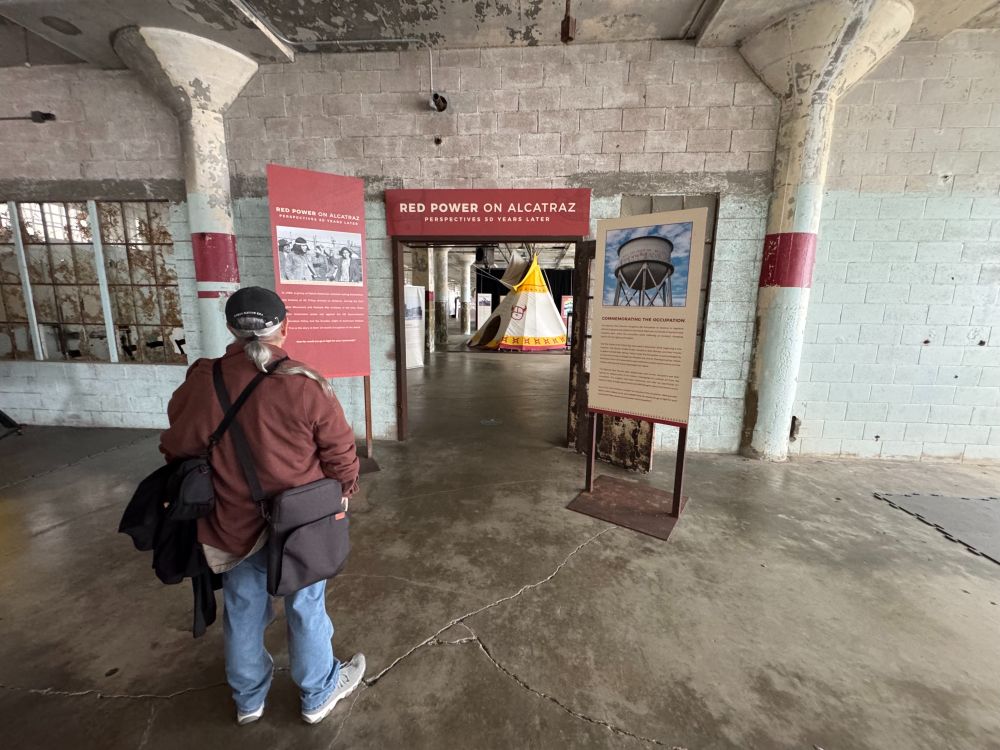 Gregg Castro from behind, approaching a door with three exhibit panels with title "RED POWER ON ALCATRAZ" on top of door. Through the door you can see a yellow and white teepee which is part of the exhibit