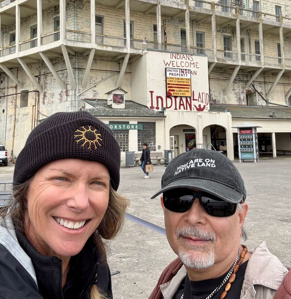 Shannon Tushingham and Gregg Castro in front of building on Alcatraz, with historic graffiti "INDIANS WELCOME UNITED INDIAN PROPERTY...INDIAN LAND" written on above, below, and top of an official sign. 