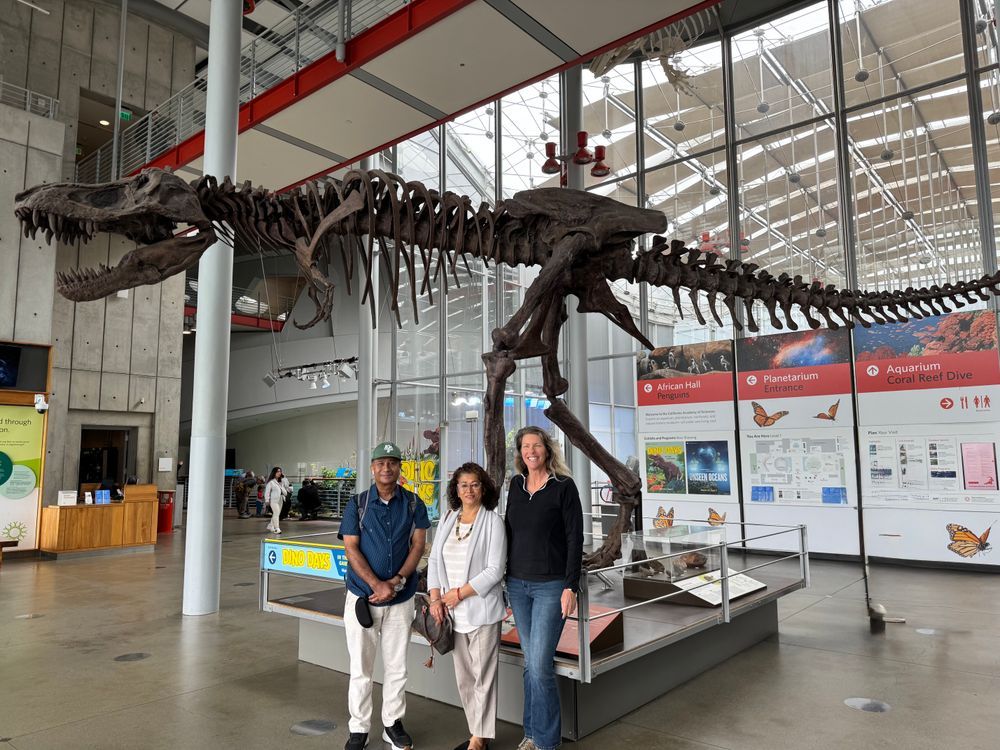 Three people standing and smiling in front of a fossil t-rex skeleton. 