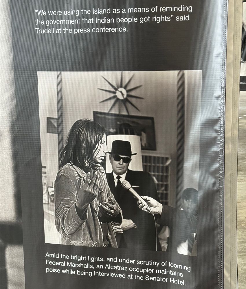 Image of Native American Alcatraz occupoier calmly speaking to a reporter, with federal marshall in black hat, suit, sunglasses observing in the background, looking like a menacing "blues brother" 