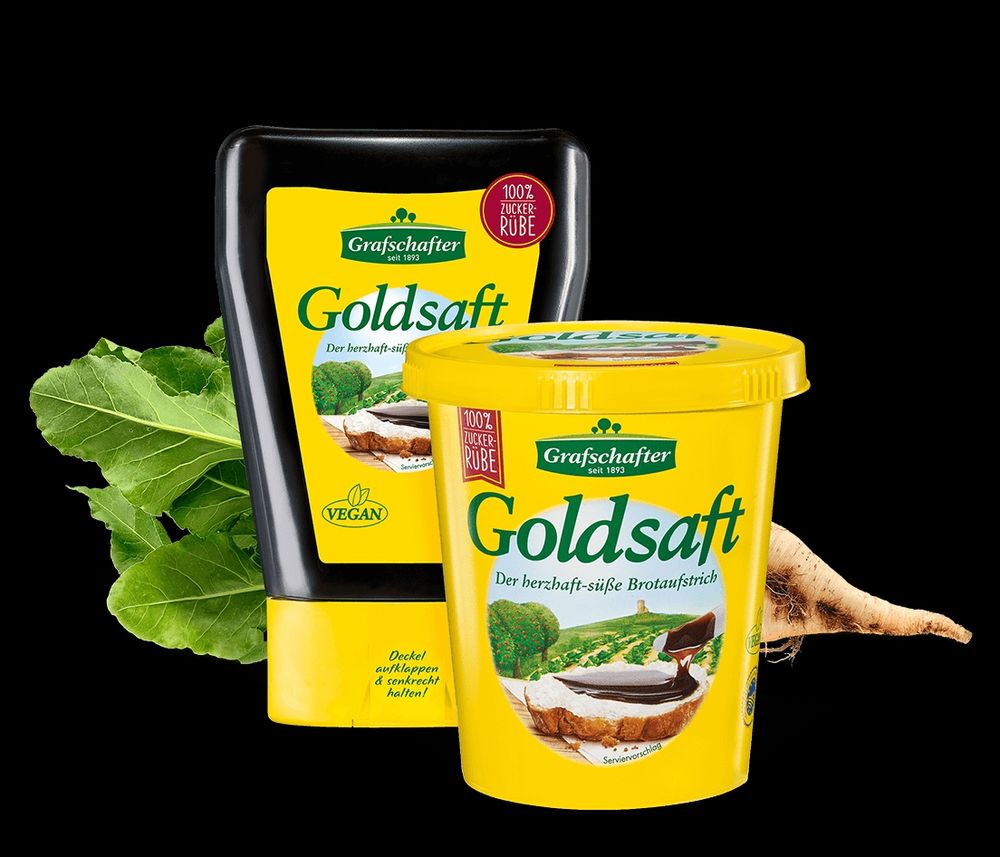 Im Vordergrund ist ein gelber Becher mit Aufdruck "Grafschafter Goldsaft, der herzhaft-süße Brotaufstrich". Leicht links dahinter ist eine Dosiertube mit der gleichen Aufschrift. Dahinter liegt eine Zuckerrübe 