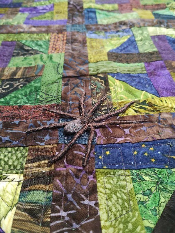 closeup of a quilt in mostly greens and browns, some blue, with a realistic (ish) embroidered spider in the middle